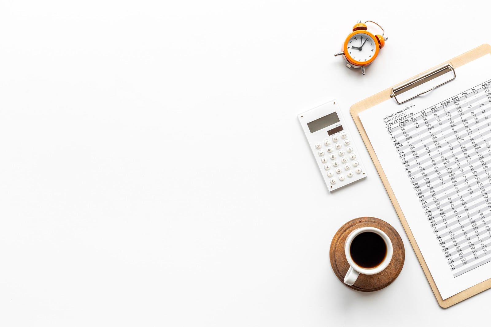 Kop kaffe, lommeregner, vækkeur og et clipboard med papirer på et hvidt skrivebord, set oppefra
